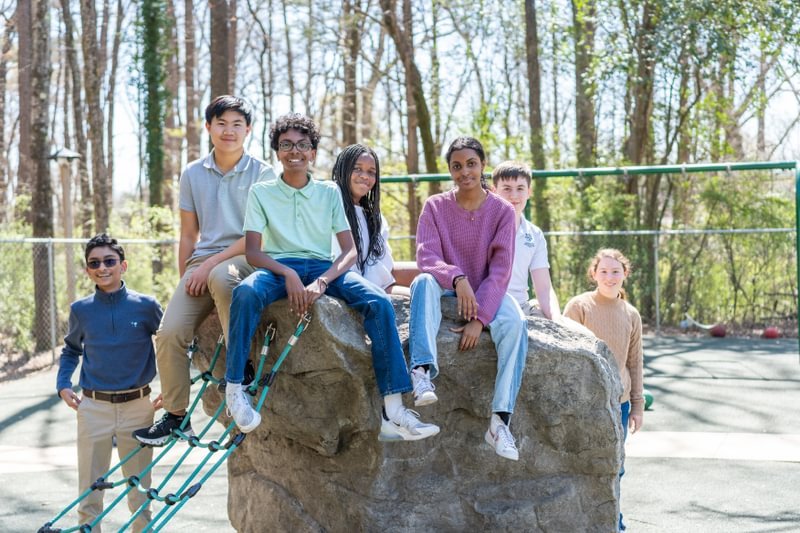 Seven students sit on and around a rock on a playground smiling.