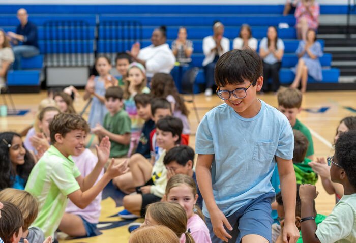 Student walks and smiles surrounded by students seated on the floor, smiling and clapping.