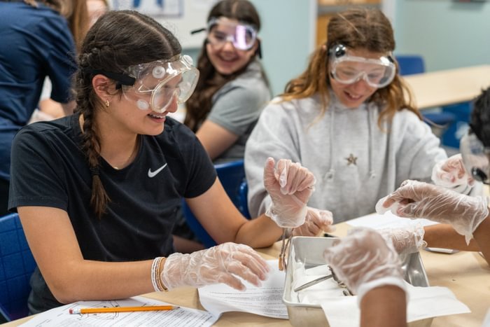 Students in safety goggles and gloves smile at project on the table below them.