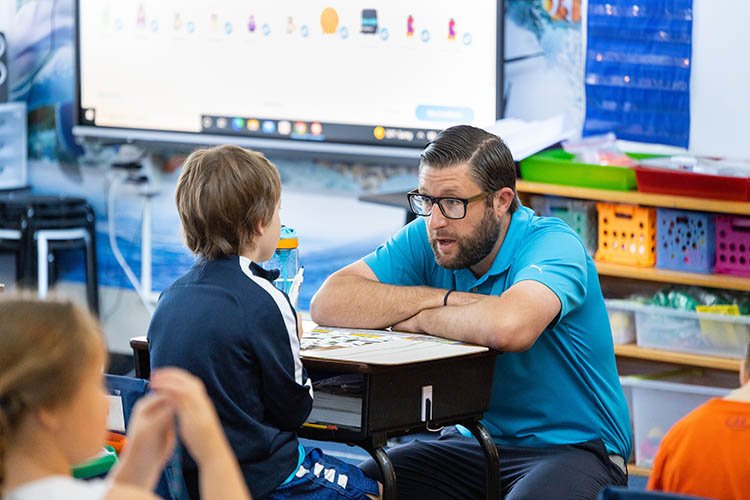 A teacher crouches down to a students desk